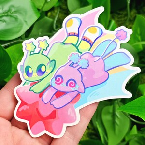May include: Two cartoon characters, one green and one pink, are flying on a pink star with a rainbow tail. The green character has large eyes and two stars on its head. The pink character has large eyes with spiral patterns and a flower on its head.
