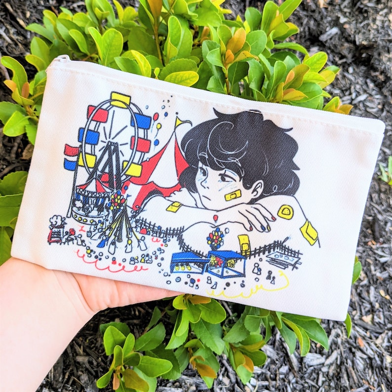 May include: White zippered pouch with a colorful illustration of a person with black hair and a Ferris wheel. The person is wearing bandages and is sitting with their arms crossed. The background is filled with colorful details.