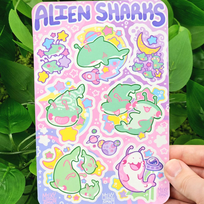 Puede incluir: Una hoja de pegatinas con el texto "ALIEN SHARKS" en la parte superior. La hoja presenta ilustraciones de tiburones alien&iacute;genas de dibujos animados en tonos verdes y rosas, con estrellas, planetas y una luna creciente sobre un fondo rosa.