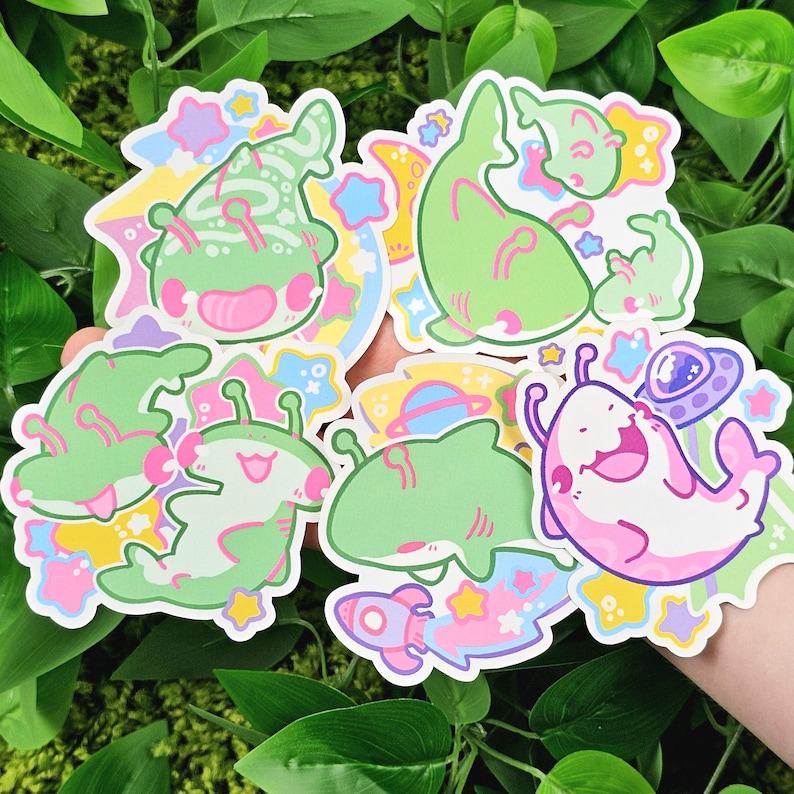 May include: A collection of colorful stickers featuring cartoon aliens and space-themed elements. The stickers showcase green, pink, and white aliens with various designs, alongside rockets, planets, and UFOs. The stickers have a playful and whimsical aesthetic.