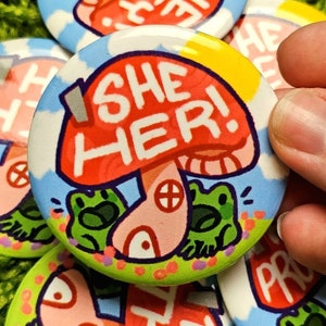 May include: A red and white mushroom with the text "SHE HER!" on it. Two green frogs are sitting on either side of the mushroom. The background is a blue sky with white clouds.