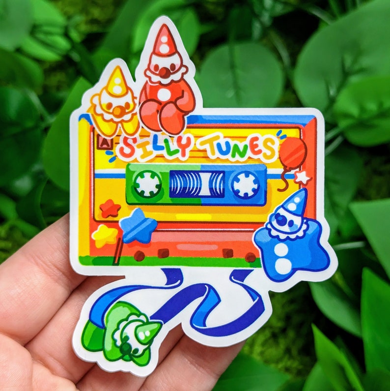 以下が含まれることがあります： 「Silly Tunes」と書かれたカラフルなカセットテープのステッカー。ステッカーには、ピエロ、星型の青いキャラクター、星型の黄色いキャラクターなど、4人の漫画のキャラクターが描かれています。