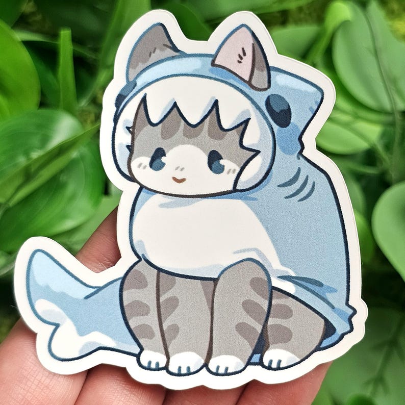 Puede incluir: Un gato gris y blanco con un disfraz de tibur&oacute;n azul. El gato est&aacute; sentado con las piernas cruzadas y tiene una expresi&oacute;n linda en su rostro.