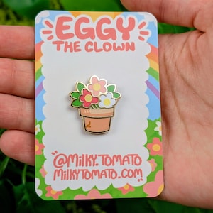 May include: An enamel pin depicting a flower pot with pink, red, and white flowers. The pin is on a card with the text "Eggy The Clown" and "@milky.tomato milkytomato.com". The background has a rainbow and floral design.