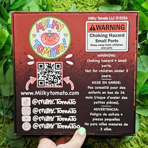May include: A red box with a cartoon tomato logo and the text "Milky Tomato" on the front. The box has a warning label about choking hazards and small parts. The box also includes a QR code and social media handles for Milky Tomato.