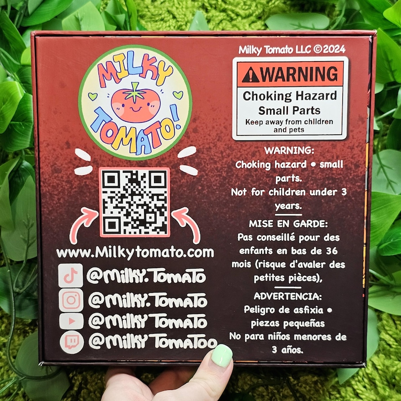 May include: A red box with a cartoon tomato logo and the text "Milky Tomato" on the front. The box has a warning label about choking hazards and small parts. The box also includes a QR code and social media handles for Milky Tomato.