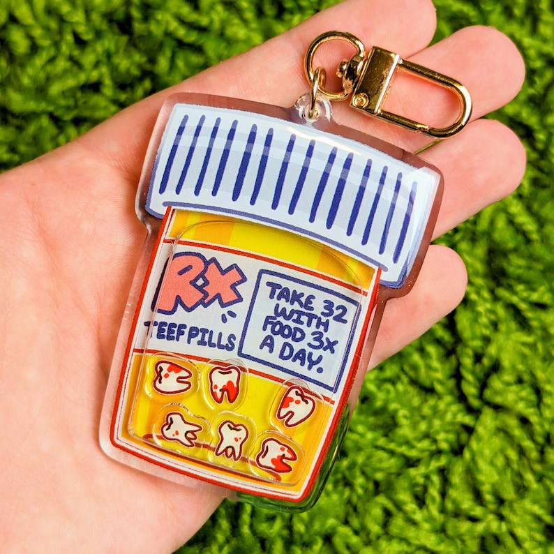 May include: A clear plastic keychain shaped like a pill bottle with a yellow label that says "Rx Teefpills Take 32 with food 3x a day." Inside the bottle are six red and white tooth-shaped candies.