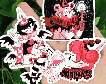 May include: A collection of four die-cut stickers featuring whimsical, gothic-inspired illustrations. The stickers showcase characters in red, black, and white, with elements like bats, ruffs, and a candle. The overall theme is a blend of fantasy and dark aesthetics.