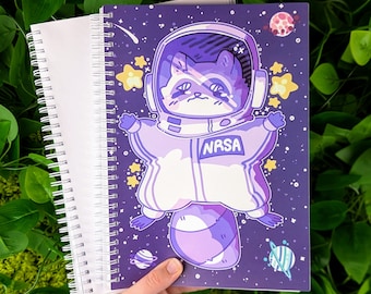 May include: A purple spiral notebook with a space-themed illustration of a raccoon wearing a space suit and the text "NRSA". The raccoon is surrounded by stars and planets.