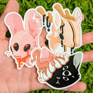 May include: Three colorful stickers featuring a pink bunny with dripping red paint, a light brown deer with black dripping paint, and a black cat with a pink tongue and sharp teeth.