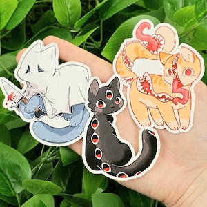 May include: A collection of cat-themed stickers. One sticker features a white cat in a ghost costume holding a bloody knife. Another shows a black cat with multiple red eyes. The third sticker is an orange cat with a tentacle tail and a mouth on its side.