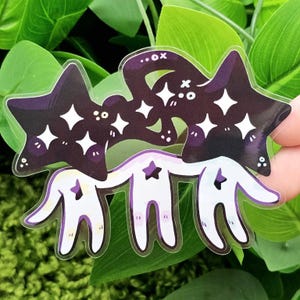 May include: A clear sticker featuring a whimsical design. Two dark purple star shapes with white diamond accents are connected to three white figures with star-shaped bodies and arms. The sticker has a glossy finish and is set against a backdrop of green leaves.