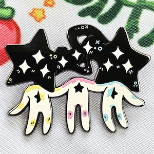 Puede incluir: Un pin de esmalte caprichoso con tres figuras estilizadas con cabezas en forma de estrella y brazos extendidos. Las cabezas de estrella negras tienen detalles de diamantes blancos. Las figuras son blancas con detalles amarillos, rosas y azules.