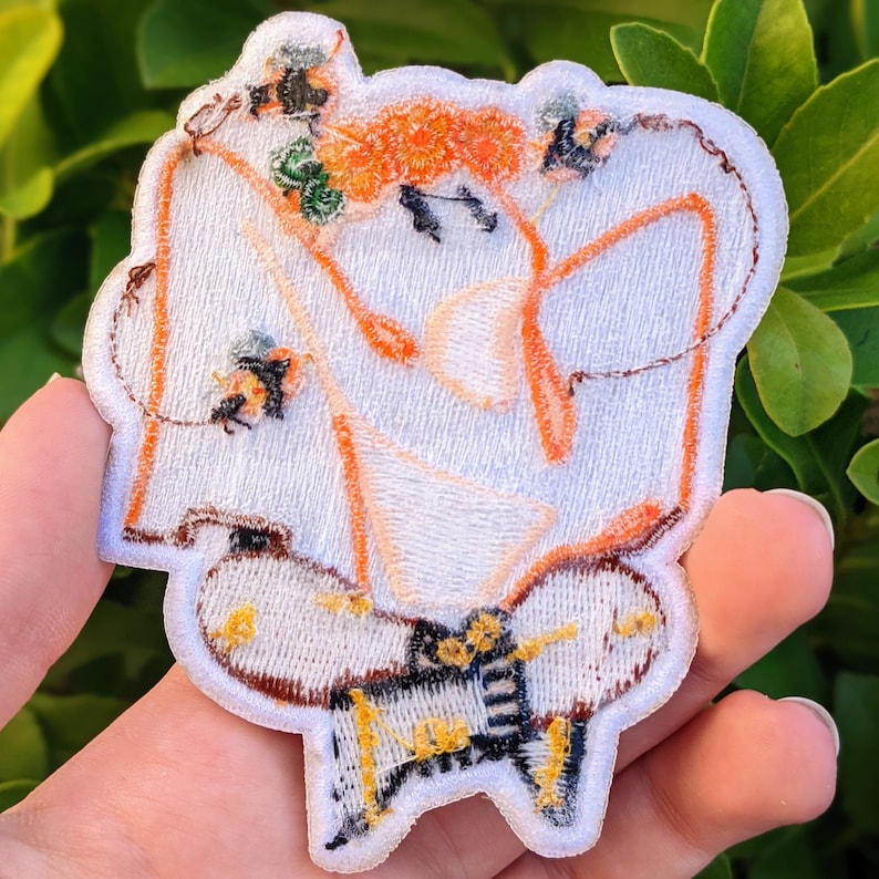 May include: White embroidered patch with a stylized figure sitting in a lotus position. The figure is outlined in orange and has small bees embroidered around it.
