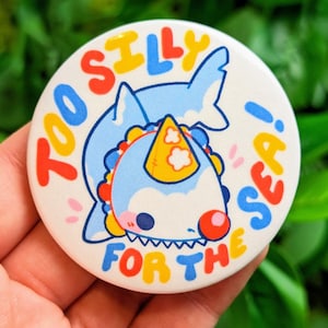 May include: A white button featuring a blue shark wearing a yellow party hat. The text on the button reads "Too silly for the sea!"