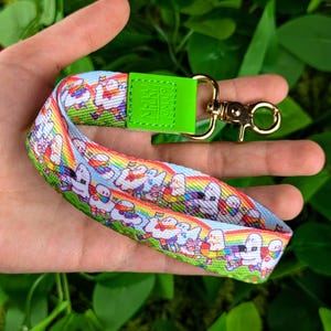 May include: A green and white lanyard with a rainbow pattern and a gold metal clasp. The lanyard features a repeating pattern of cartoon ghosts holding rainbow flags. The text "MELODY" is printed on the green strap.