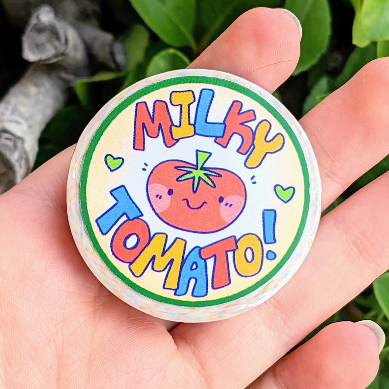 May include: A round sticker with a yellow background and green border. The sticker features a cartoon tomato with a smiling face and the text "MILKY TOMATO!" in colourful letters.