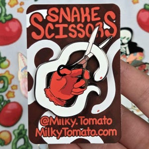 May include: A metal enamel pin featuring a red hand holding silver scissors, surrounded by a white snake. The pin is attached to a rectangular backing card with the text SNAKE SCISSORS, @Milky.Tomato, and MilkyTomato.com printed in orange.