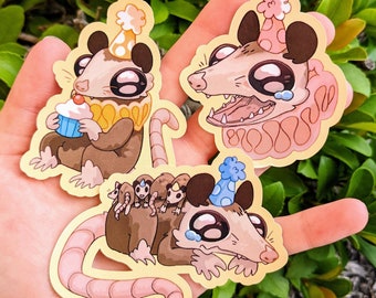 Könnte beinhalten: Drei niedliche Cartoon-Opossums, die Partyhüte tragen. Ein Opossum hält einen Cupcake, ein anderes hält ein kleineres Opossum und das dritte Opossum weint. Die Opossums befinden sich alle auf einem weißen Hintergrund.