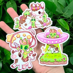 May include: A collection of colorful stickers featuring cartoon aliens and cows. One sticker shows an alien riding a cow with a lasso and a gun. Another shows a cow being beamed up into a pink UFO. The stickers have a whimsical, playful design.