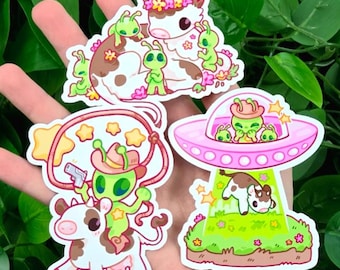May include: A collection of colorful stickers featuring cartoon aliens and cows. One sticker shows an alien riding a cow with a lasso and a gun. Another shows a cow being beamed up into a pink UFO. The stickers have a whimsical, playful design.