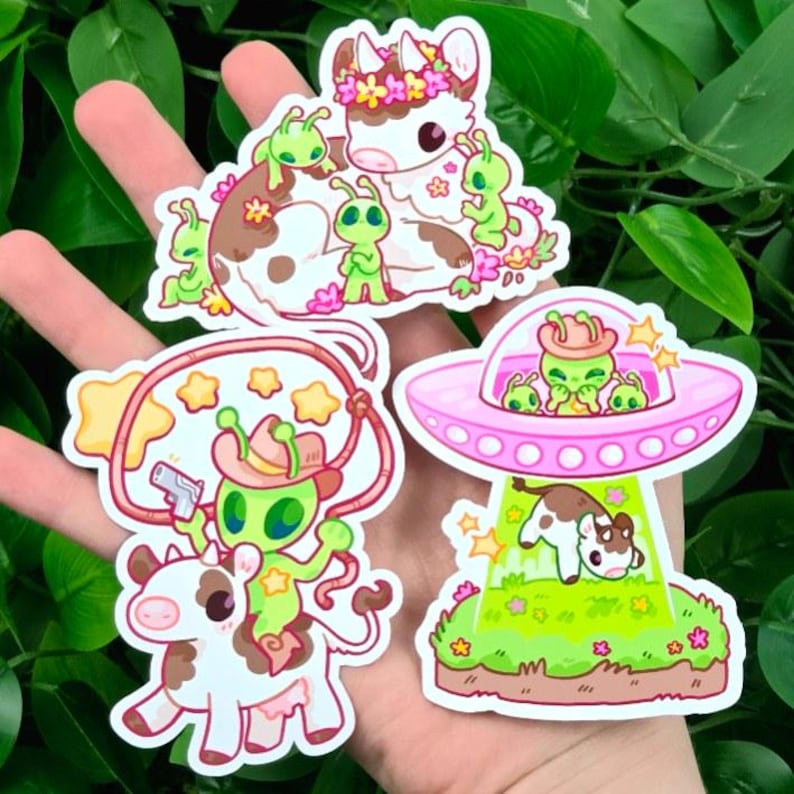 May include: A collection of colorful stickers featuring cartoon aliens and cows. One sticker shows an alien riding a cow with a lasso and a gun. Another shows a cow being beamed up into a pink UFO. The stickers have a whimsical, playful design.