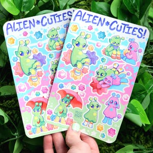 May include: Two sticker sheets with a pink and blue background featuring cute alien characters with rainbow accents. The sticker sheets are titled "Alien Cutie's!"