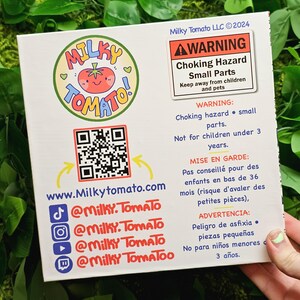 May include: A colourful cartoon tomato logo with the word "Milky Tomato" in a circle. The logo is on a white background with a QR code, a website address, and social media handles. The image also includes a warning label about choking hazards for children under 3 years old.
