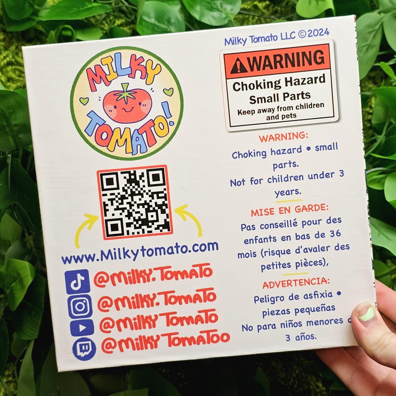 May include: A colourful cartoon tomato logo with the word "Milky Tomato" in a circle. The logo is on a white background with a QR code, a website address, and social media handles. The image also includes a warning label about choking hazards for children under 3 years old.
