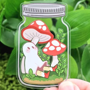 Puede incluir: Una pegatina transparente con un fantasma de dibujos animados sentado en un tarro con dos setas rojas y blancas y follaje verde. El tarro tiene una tapa plateada. El fantasma es blanco con ojos negros y lleva zapatos rojos y amarillos.