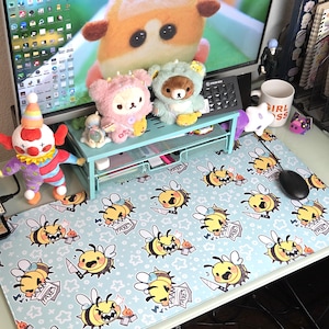 Pode incluir: Tapete de rato azul claro com um padr&atilde;o repetido de abelhas de desenho animado segurando facas e formul&aacute;rios fiscais. A mesa tamb&eacute;m tem um monitor, uma boneca de palha&ccedil;o e v&aacute;rios brinquedos de pel&uacute;cia.