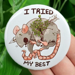 May include: A white button with the text "I TRIED MY BEST" encircling a cartoon illustration of a gray rat pierced by an arrow and adorned with green foliage. The rat has pink ears and a pink tail.