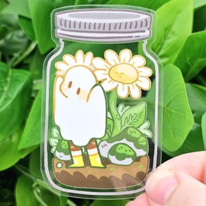 Puede incluir: Una pegatina transparente con un fantasma de dibujos animados dentro de un tarro con margaritas y vegetación. El fantasma tiene un cuerpo blanco, ojos marrones y botas amarillas. El tarro tiene una tapa plateada y se asienta sobre una base marrón.