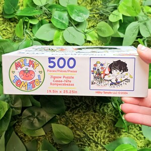 May include: A 500-piece jigsaw puzzle box featuring a cartoon tomato illustration and the words "Milky Tomato!" in a pink and white circle. The box also includes a colourful illustration of a Ferris wheel and a person sitting on a bench. The puzzle measures 19.5 inches by 25.25 inches.