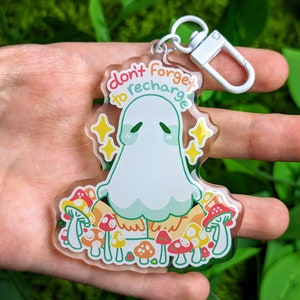 May include: A clear acrylic keychain featuring a cartoon ghost with the text "don't forget to recharge" in a rainbow gradient. The ghost is surrounded by colorful mushrooms.