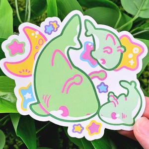 May include: A die-cut sticker featuring a whimsical design of green cartoon creatures with pink accents, surrounded by yellow moons and stars with pink, blue, and yellow details. The sticker has a white border and is held against a background of green leaves.