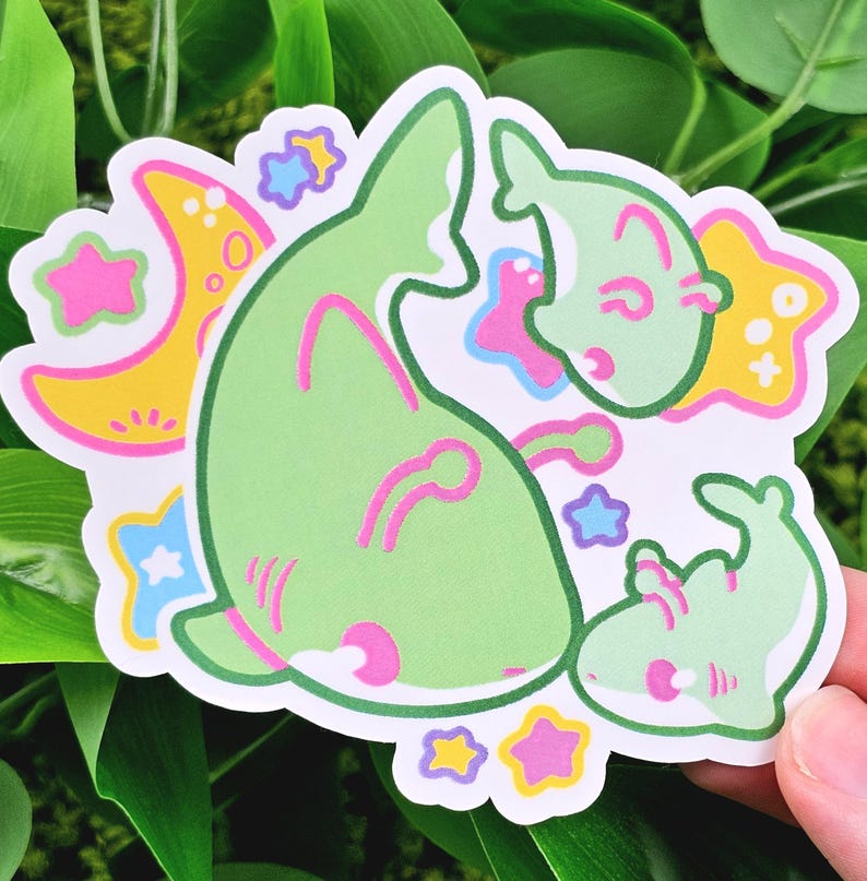 May include: A die-cut sticker featuring a whimsical design of green cartoon creatures with pink accents, surrounded by yellow moons and stars with pink, blue, and yellow details. The sticker has a white border and is held against a background of green leaves.