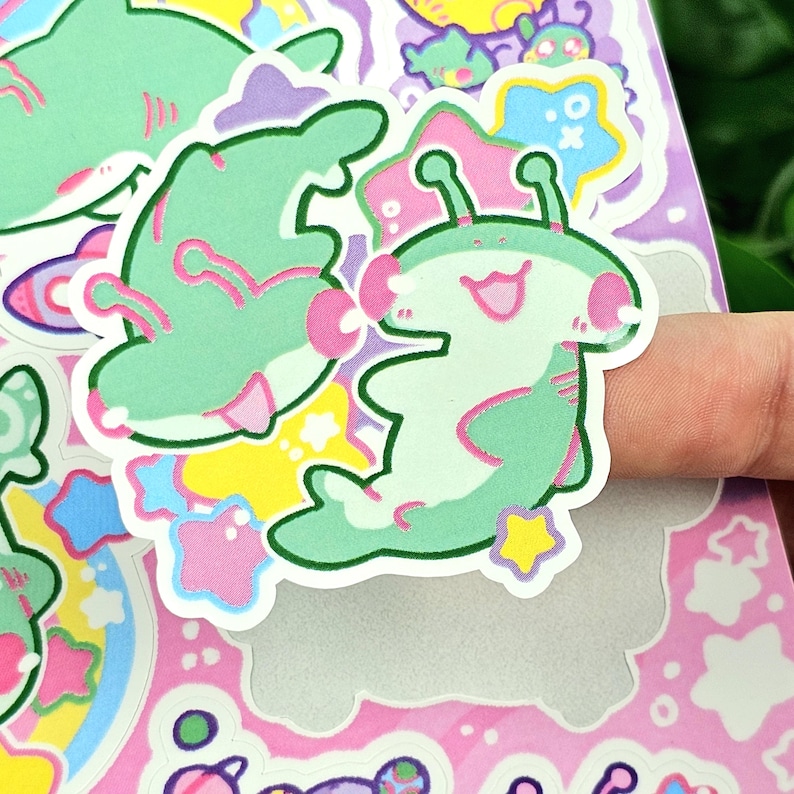 Puede incluir: Una hoja de pegatinas coloridas con extraterrestres y estrellas de dibujos animados. Las pegatinas tienen una paleta de colores pastel de verde, rosa, amarillo y azul. Las pegatinas de extraterrestres tienen una cara sonriente y antenas.