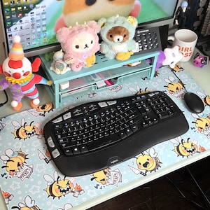 Pode incluir: Um teclado e um rato ergon&oacute;micos pretos est&atilde;o sobre um tapete de secret&aacute;ria azul claro com um padr&atilde;o de abelhas de desenho animado. A secret&aacute;ria tamb&eacute;m apresenta brinquedos de pel&uacute;cia, uma boneca palha&ccedil;o e um monitor.