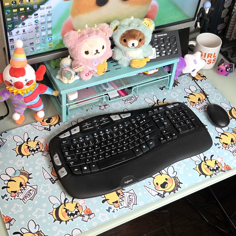 Pode incluir: Um teclado e um rato ergon&oacute;micos pretos est&atilde;o sobre um tapete de secret&aacute;ria azul claro com um padr&atilde;o de abelhas de desenho animado. A secret&aacute;ria tamb&eacute;m apresenta brinquedos de pel&uacute;cia, uma boneca palha&ccedil;o e um monitor.