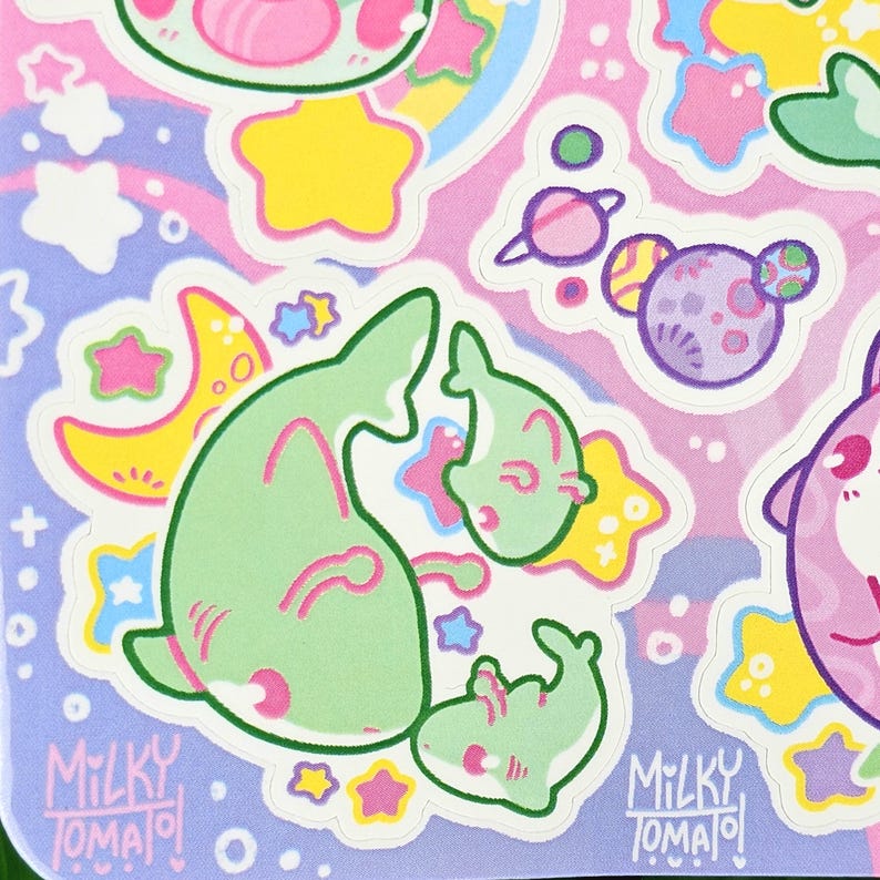 Puede incluir: Una hoja de pegatinas coloridas con ilustraciones de estilo caricatura. Los dise&ntilde;os incluyen criaturas verdes con forma de tibur&oacute;n, planetas, estrellas y lunas en colores pastel. El texto "Milky Tomato" est&aacute; impreso en la parte inferior derecha e izquierda.