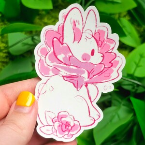 Puede incluir: Un conejito blanco con orejas rosas y una flor rosa alrededor del cuello. El conejito est&aacute; sentado en una flor rosa con hojas verdes.