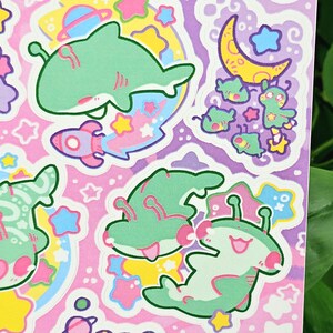 Puede incluir: Una hoja de pegatinas coloridas con tiburones, extraterrestres, cohetes, estrellas y planetas de dibujos animados. Las pegatinas tienen una paleta de colores pastel rosa, verde, amarillo y azul. Los dise&ntilde;os son caprichosos y juguetones.
