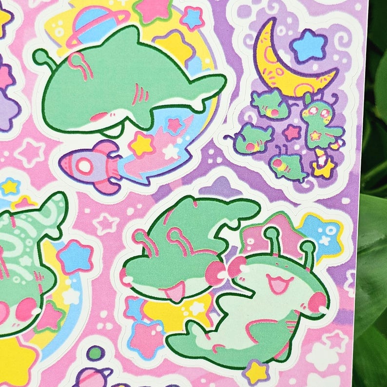 Puede incluir: Una hoja de pegatinas coloridas con tiburones, extraterrestres, cohetes, estrellas y planetas de dibujos animados. Las pegatinas tienen una paleta de colores pastel rosa, verde, amarillo y azul. Los dise&ntilde;os son caprichosos y juguetones.