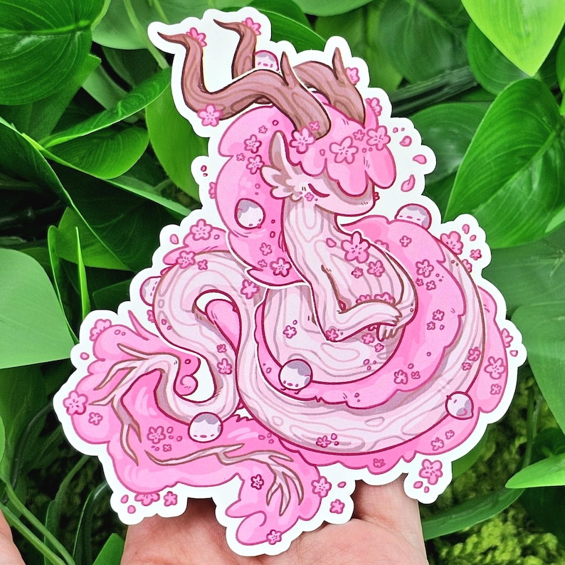 May include: A pink and white dragon-like creature with antlers and flowers. The creature is curled up in a sitting position with its tail wrapped around its body. The creature is covered in pink flowers and has a white face with closed eyes.