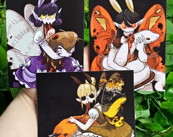 May include: Three illustrated cards featuring a dark background with a fairy-like creature with wings and a bloody mouth holding a small animal. The first card features a creature with purple wings and a rat, the second card features a creature with orange wings and a white snake, and the third card features a creature with yellow wings and a fox.