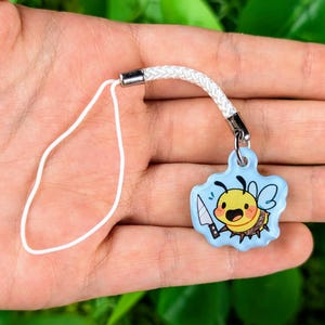 May include: A white phone charm with a blue and yellow cartoon bee holding a knife. The bee has a smiling face and is wearing a yellow and black striped body.