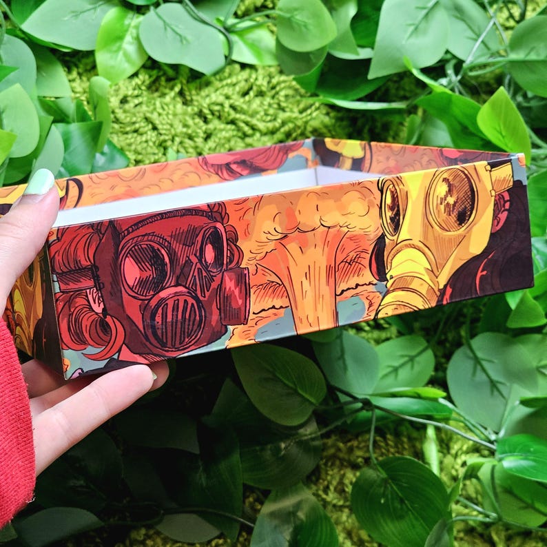 May include: A rectangular box with a colorful design featuring three gas masks and explosions. The box is made of cardboard and has a white interior.