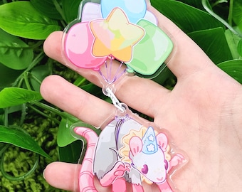 May include: A pink, gray, and white opossum wearing a party hat and holding a string of colorful balloons. The opossum is holding the balloons with its front paws. The balloons are a star, a heart, and two round balloons.