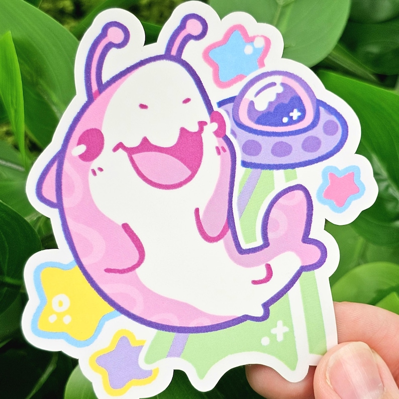 May include: A sticker featuring a cartoon alien creature in pink and white, with a large smile and purple outlines. The alien is accompanied by a purple and blue UFO, stars, and green and white beams. The sticker is held by a hand.